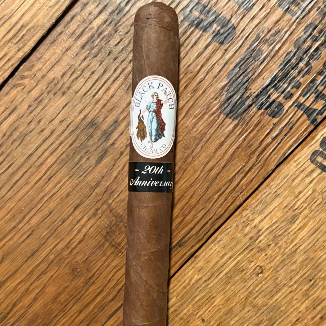 Brown cigar with Black Patch Cigar Co. band and a '20th Anniversary' label on a wooden surface.